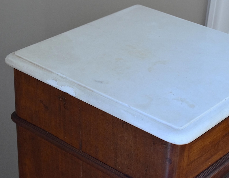 May include: A close-up of a wooden cabinet with a white marble top. The cabinet is made of dark brown wood with a curved design. The marble top has a slightly rounded edge.