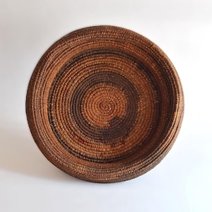 Puede incluir: Una cesta grande, redonda y tejida con un esquema de color marrón y negro. La cesta está hecha de fibras naturales y tiene una forma circular profunda.