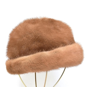 May include: A vintage brown fur hat with a rounded crown and a wide, rolled brim. The hat is displayed on a gold-colored stand against a white background. The fur appears soft and plush.