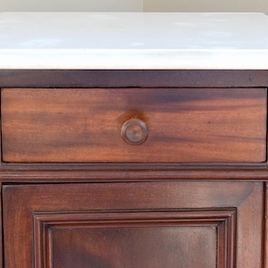 May include: A small, antique wooden cabinet with a white marble top. The cabinet has a single drawer with a round wooden knob and a lower door. The wood has a rich, dark brown finish.