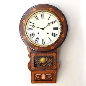 Puede incluir: Un reloj de pared con marco de madera circular con patrones circulares incrustados y esfera blanca con números romanos negros. Debajo de la esfera hay un panel de vidrio rectangular que muestra un emblema de águila dorada y una sección inferior.