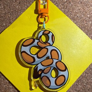 Op de afbeelding: Een heldere acryl sleutelhanger met een cartoon slang ontwerp. De slang is lichtblauw met bruine vlekken en een donkerbruine kop. Hij hangt aan een oranje sluiting die is bevestigd aan een geel ruitvormig papier.