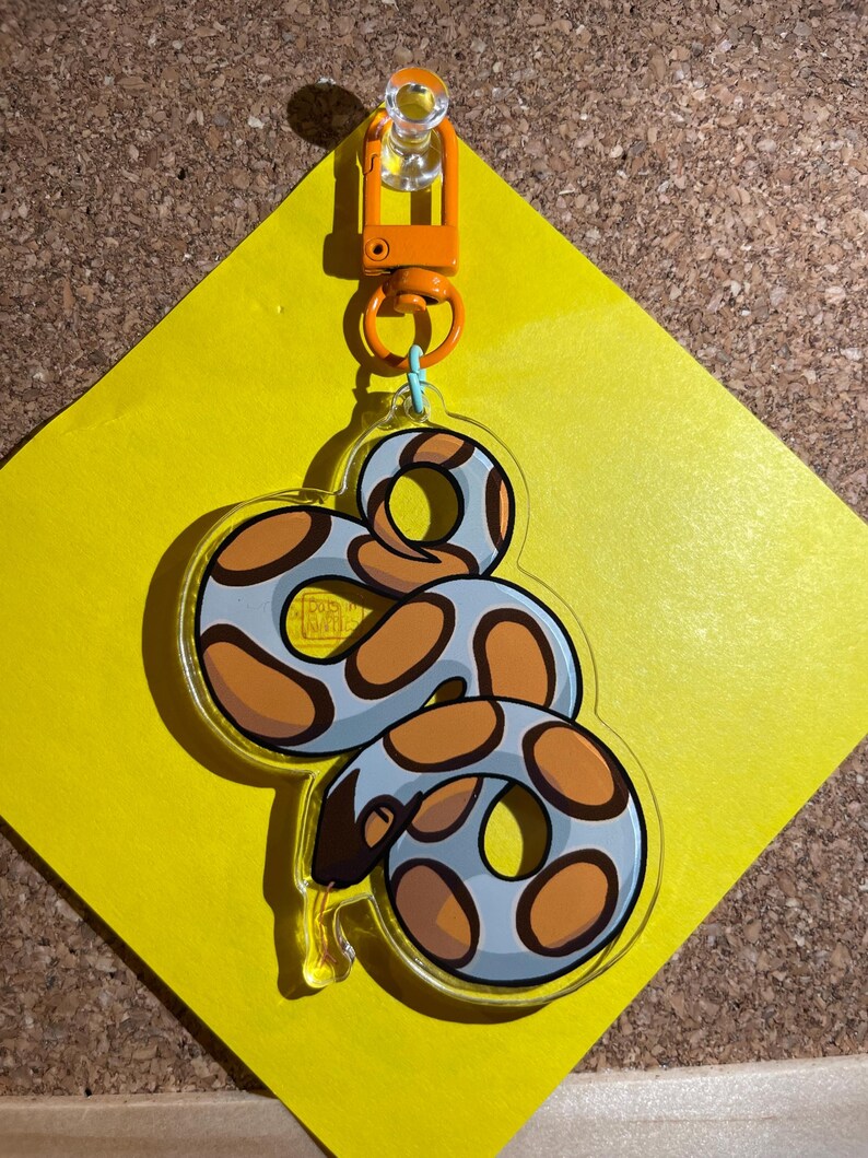 Op de afbeelding: Een heldere acryl sleutelhanger met een cartoon slang ontwerp. De slang is lichtblauw met bruine vlekken en een donkerbruine kop. Hij hangt aan een oranje sluiting die is bevestigd aan een geel ruitvormig papier.
