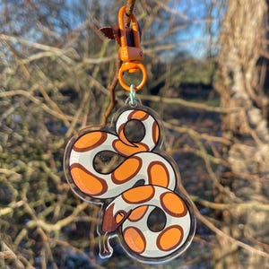 Peut inclure: Un porte-clés en acrylique transparent avec un motif de serpent de dessin animé. Le serpent a un corps blanc avec des marques orange et marron. Le porte-clés est attaché à un fermoir orange et un petit anneau vert.