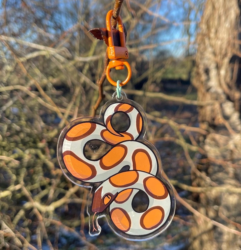 Op de afbeelding: Een heldere acryl sleutelhanger met een cartoon slang ontwerp. De slang heeft een wit lichaam met oranje en bruine markeringen. De sleutelhanger is bevestigd aan een oranje sluiting en een kleine groene ring.