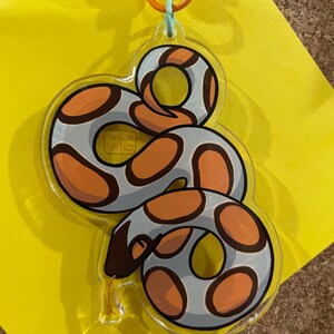 Op de afbeelding: Een heldere acryl sleutelhanger met een cartoon slang ontwerp. De slang heeft een grijze body met oranje en bruine vlekken. De sleutelhanger heeft een oranje sluiting en een lichtblauwe ring.