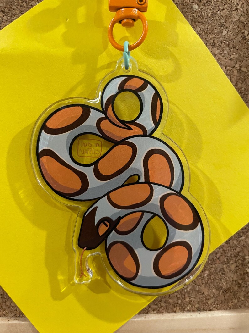 Op de afbeelding: Een heldere acryl sleutelhanger met een cartoon slang ontwerp. De slang heeft een grijze body met oranje en bruine vlekken. De sleutelhanger heeft een oranje sluiting en een lichtblauwe ring.