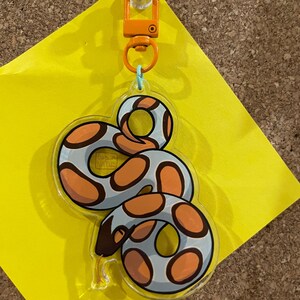 Op de afbeelding: Een heldere acryl sleutelhanger met een cartoon slang ontwerp. De slang heeft een lichtblauw lichaam met oranje en bruine vlekken. Het heeft een oranje sluiting en een lichtblauwe ring. De sleutelhanger is bevestigd aan een gele plakbriefje.