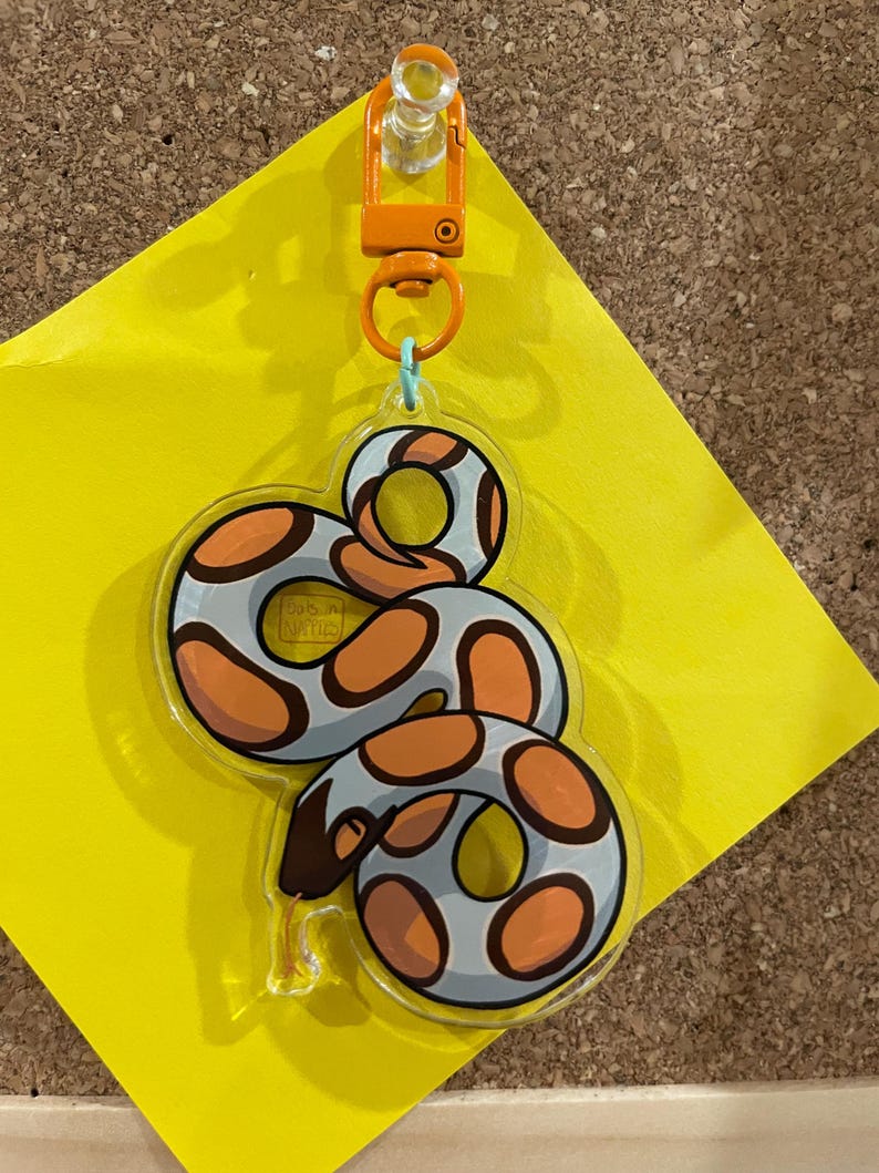 Op de afbeelding: Een heldere acryl sleutelhanger met een cartoon slang ontwerp. De slang heeft een lichtblauw lichaam met oranje en bruine vlekken. Het heeft een oranje sluiting en een lichtblauwe ring. De sleutelhanger is bevestigd aan een gele plakbriefje.