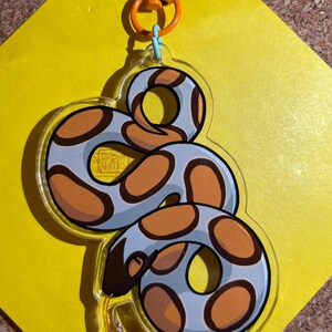 Op de afbeelding: Een heldere acryl sleutelhanger met een cartoon slang ontwerp. De slang heeft een lichtblauw lichaam met oranje en bruine vlekken. De sleutelhanger heeft een oranje sluiting en een lichtblauwe ring.