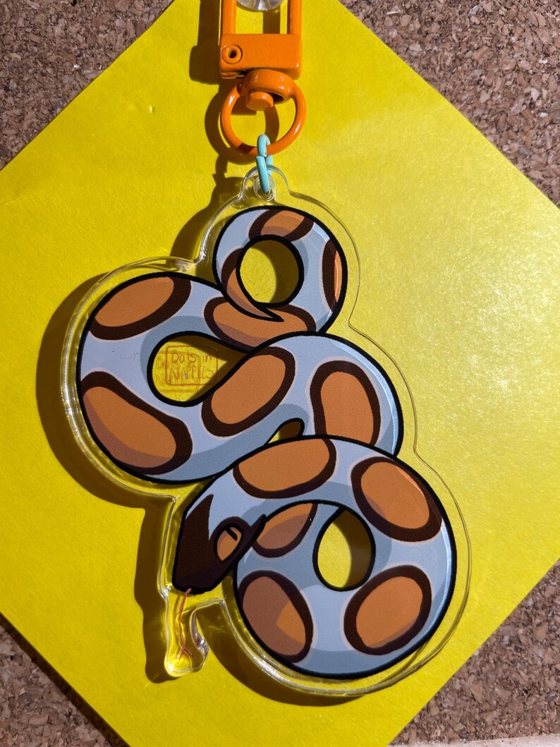 Op de afbeelding: Een heldere acryl sleutelhanger met een cartoon slang ontwerp. De slang heeft een lichtblauw lichaam met oranje en bruine vlekken. De sleutelhanger heeft een oranje sluiting en een lichtblauwe ring.