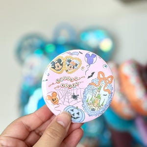 Puede incluir: Un botón redondo con fondo rosa claro que presenta una ilustración colorida de Mickey y Minnie Mouse, un castillo, telarañas, calabazas y globos. El texto "SPOOKY VIBES" está impreso en el centro con una tipografía divertida.