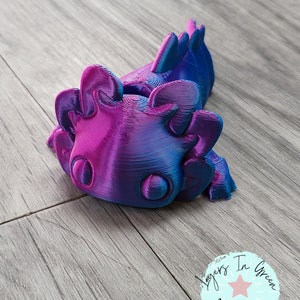 May include: A 3D printed axolotl figurine with a blue and purple ombre finish. The figurine is lying on its side with its tail curled up. The figurine has a detailed design with a textured surface.