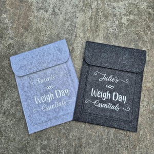 May include: Two felt pouches with white embroidery. The pouch on the left is light grey and says "Karen's Weigh Day Essentials". The pouch on the right is dark grey and says "Julie's Weigh Day Essentials".