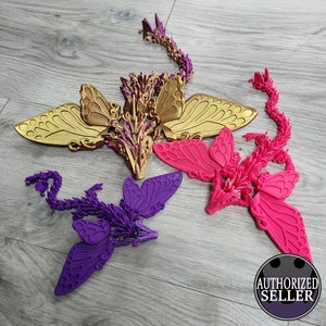 May include: Three decorative dragon sculptures with butterfly wings in gold, hot pink, and purple. Each dragon has a detailed body and a long, segmented tail. The wings are large and detailed. The sculptures are displayed on a light wood surface.