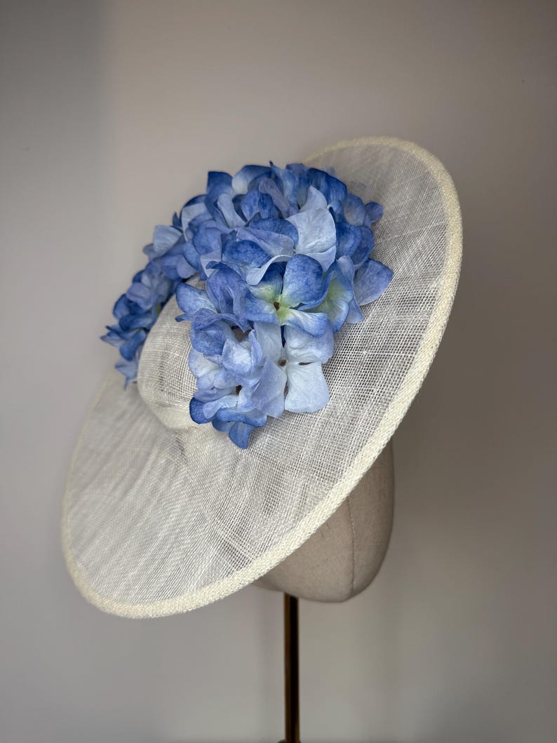 May include: A cream-colored hat with a wide brim, adorned with a cluster of blue hydrangea flowers. The hat has a woven texture and a rounded crown, suitable for special occasions.