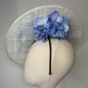 May include: A light-colored, wide-brimmed hat with a cluster of blue hydrangea flowers. The hat is displayed on a mannequin head. The brim has a woven texture. A dark brown ribbon is attached to the hat.