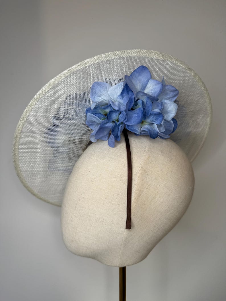 May include: A light-colored, wide-brimmed hat with a cluster of blue hydrangea flowers. The hat is displayed on a mannequin head. The brim has a woven texture. A dark brown ribbon is attached to the hat.