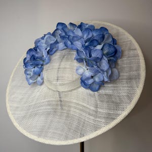 May include: A wide-brimmed, cream-colored hat with a woven texture. The hat is adorned with a garland of blue hydrangea flowers. The hat is displayed on a stand, showcasing its elegant design, suitable for special occasions.