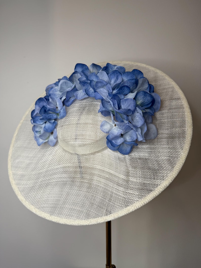 May include: A wide-brimmed, cream-colored hat with a woven texture. The hat is adorned with a garland of blue hydrangea flowers. The hat is displayed on a stand, showcasing its elegant design, suitable for special occasions.