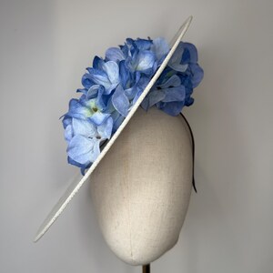 May include: A white hat with a wide brim, adorned with a cluster of light blue and white artificial hydrangea flowers. The hat is mounted on a mannequin head, showcasing its design and shape. The hat is a fashion accessory.
