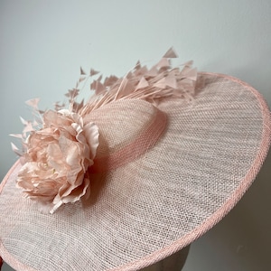 May include: A pale pink wide-brimmed hat with a woven texture, a large fabric flower, and feather detailing. The hat has a pink trim and is displayed on a mannequin head. The hat is suitable for special occasions.