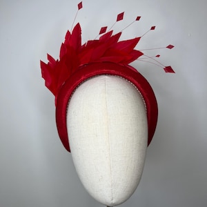 Könnte beinhalten: Auffälliger roter Kopfschmuck mit einem satinbezogenen, breiten Stirnband. Der Kopfschmuck ist mit roten Federn und rautenförmigen Akzenten auf dünnen Stielen verziert, was ein elegantes Design ergibt. Der Kopfschmuck wird auf einem Mannequinkopf präsentiert.