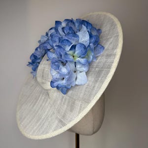 May include: A cream-colored hat with a wide brim, adorned with a cluster of blue hydrangea flowers. The hat has a woven texture and a rounded crown, suitable for special occasions.