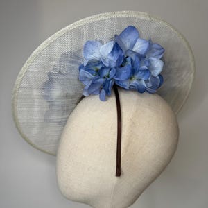 May include: A light-colored, wide-brimmed hat with a cluster of blue hydrangea flowers. The hat is displayed on a mannequin head, showcasing its shape and design. The brim has a woven texture, and a dark brown ribbon is attached.