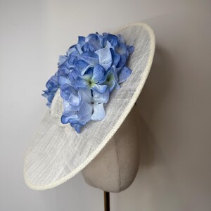 May include: A wide-brimmed, off-white hat adorned with a cluster of light blue and periwinkle hydrangea flowers. The hat is displayed on a mannequin head, showcasing its shape and design. The brim has a subtle texture.
