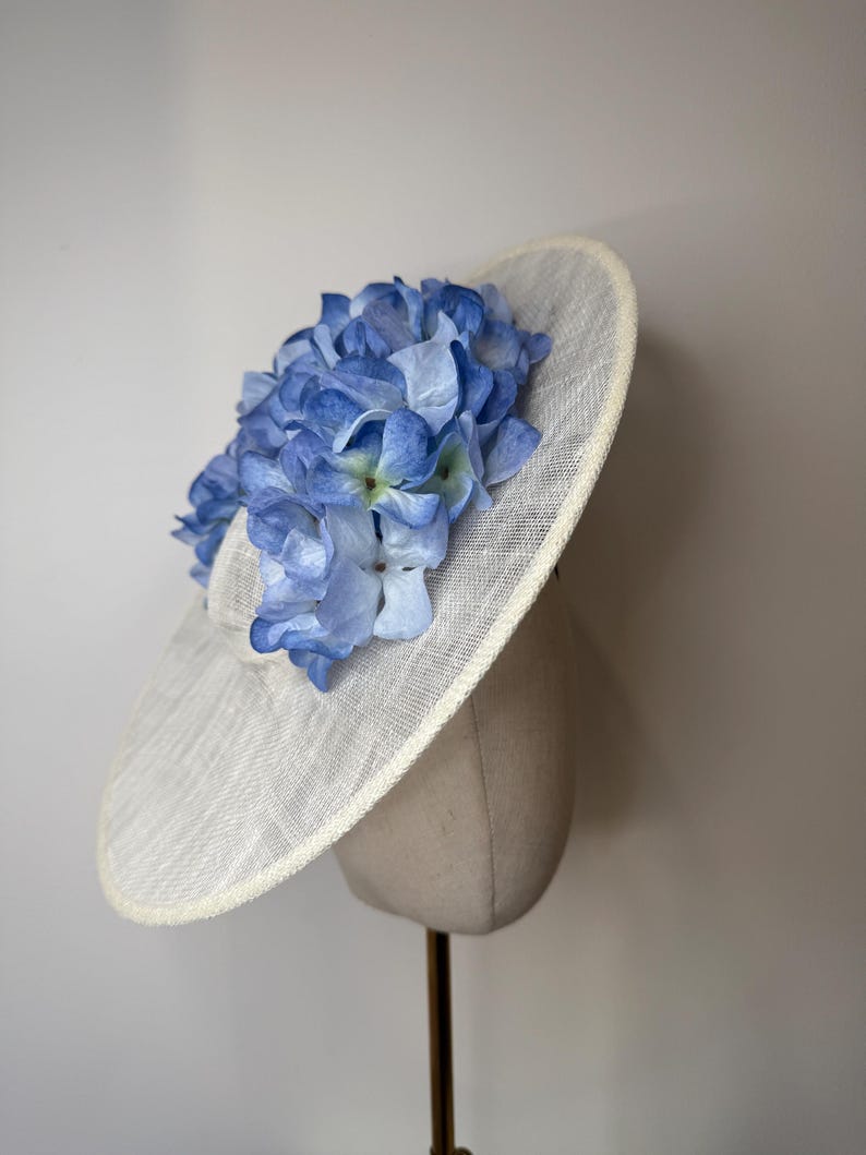 May include: A wide-brimmed, off-white hat adorned with a cluster of light blue and periwinkle hydrangea flowers. The hat is displayed on a mannequin head, showcasing its shape and design. The brim has a subtle texture.