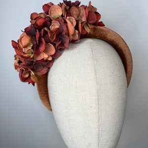 May include: A brown headband adorned with a cluster of faux flowers in shades of burgundy, rust, and tan. The flowers are densely packed, creating a full, textured appearance. The headband itself is a solid, muted brown.