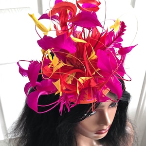May include: A fascinator headpiece featuring a vibrant arrangement of hot pink, bright orange, and yellow feathers. The feathers are layered and fanned out in a circular pattern, accented by thin, curved wire stems that extend upwards from the base.