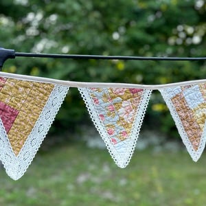 May include: A fabric bunting banner featuring triangular flags made from quilted floral fabric in shades of mustard yellow, pink, and light blue. Each triangular flag is edged with white lace trim and attached to a light beige fabric ribbon.