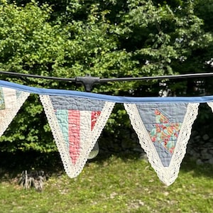 May include: A string of triangular fabric bunting flags featuring a patchwork design with various floral and striped patterns in shades of red, blue, and cream. Each flag is edged with white lace trim and attached to a blue fabric header.