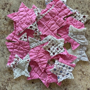 May include: A collection of fabric star-shaped cutouts featuring a mix of solid pink cotton and various white cotton fabrics with small floral and geometric patterns. The stars have raw, pinked edges and a quilted texture throughout the fabric surface.