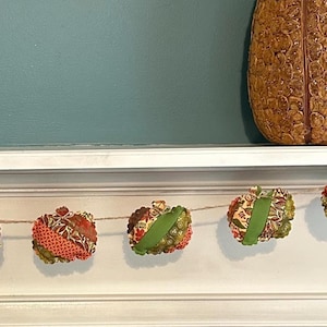 May include: A garland of fabric pumpkins in shades of orange, green, and brown, strung on twine. Each pumpkin has a green ribbon stem. The pumpkins are displayed on a white shelf with a teal wall in the background.