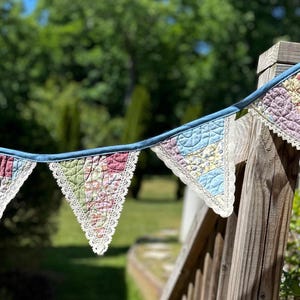 May include: A fabric bunting banner featuring four triangular quilted pennants in various floral and geometric patterns of pink, green, yellow, and blue. Each pennant is edged with white lace trim and attached to a solid blue fabric header tape.