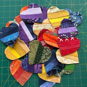 May include: A collection of heart-shaped fabric patches, each constructed from multiple horizontal strips of cotton fabric in various colors and patterns, including shades of red, orange, yellow, green, blue, purple, and floral or geometric prints.