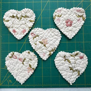 May include: Five heart-shaped fabric cutouts made of white quilted material. Each heart features embroidered floral patterns in shades of pink and olive green. The edges of the fabric hearts are scalloped, showcasing a textured, stitched surface.