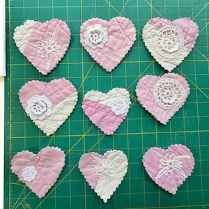 May include: Nine heart-shaped fabric ornaments arranged in a grid. Each heart is crafted from quilted pink and cream fabric with scalloped edges. The hearts are decorated with various white crocheted lace appliques, including floral and star patterns.
