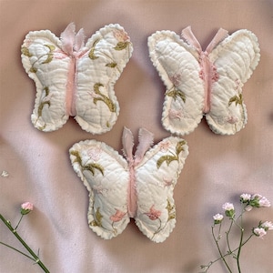 May include: Three quilted butterfly ornaments made of off-white fabric with pink and gold floral embroidery. Each butterfly features a central pink fabric strip and small frayed fabric antennae at the top, showcasing a soft, textured surface.