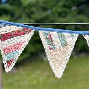 May include: A fabric bunting banner featuring triangular pennants made from quilted patchwork fabric in various floral and striped patterns. Each pennant is edged with white lace trim and attached to a solid blue fabric header strip.