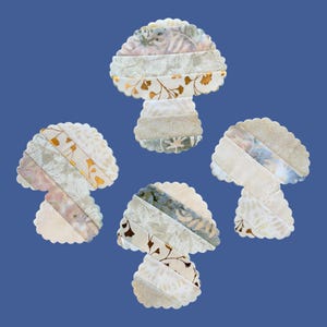 May include: Four mushroom-shaped fabric appliques featuring a scalloped edge. Each piece is constructed from diagonal strips of fabric in shades of cream, beige, and soft grey, with some sections displaying metallic gold leaf botanical patterns.