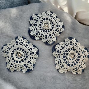 May include: Three white crochet flower appliqués with a denim blue backing and a pearl bead center. The handmade floral embellishments are arranged on a gray fabric surface. These decorative elements can be used for various craft projects.