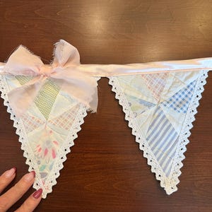 May include: A fabric bunting banner featuring two triangular pennants made of quilted patchwork fabric in pastel shades of blue, pink, and green. The edges are trimmed with white lace, and a light pink ribbon bow is attached to the top corner.