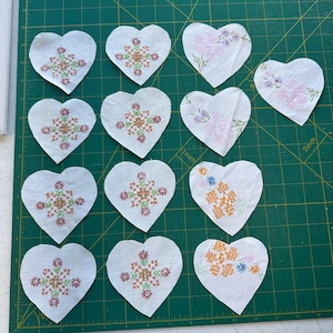 May include: Eleven white fabric hearts arranged in a grid, each featuring cross-stitched floral patterns. Eight hearts display symmetrical geometric flower designs in pink and brown, while three hearts feature asymmetrical floral clusters in pink, blue, and orange.