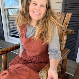 May include: A rust-colored linen apron dress with a square neckline and a gathered waist. The dress is worn over a gray and white striped long-sleeved shirt. The person is sitting in a wooden rocking chair on a porch.