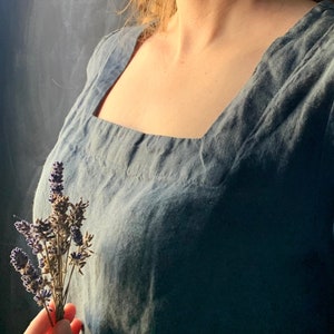 May include: A woman wearing a blue linen dress with a square neckline, holding a bouquet of dried lavender.