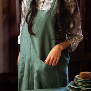 May include: A woman wearing a green linen apron with a large pocket. She is standing in front of a table with a stack of plates and a piece of cake on top.